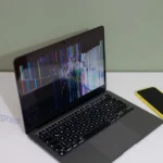 A cracked laptop screen on a messy student desk with a small jar of coins nearby