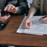 A printed tax form next to an empty coffee mug and a notebook showing math calculations on a wooden table outdoors