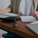 A close-up shot of a young adult's hands using a calculator next to a laptop displaying tax forms and tax documents spread out on the desk.
