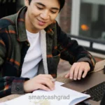 A young adult student focused on typing on a laptop at a small table with textbooks and a coffee cup nearby.